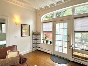 A room with a brown leather sofa, a framed artwork, a black metal stand with potted plants, and a large window with a view of greenery. - “2-bedroom apartment in a historic carriage house with garden” (Lambertville)