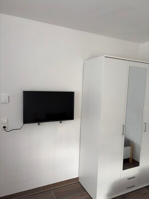 Ein Zimmer mit einem flachbildfernseher an der Wand, einem weißen Kleiderschrank mit Spiegel und einem Holzboden.