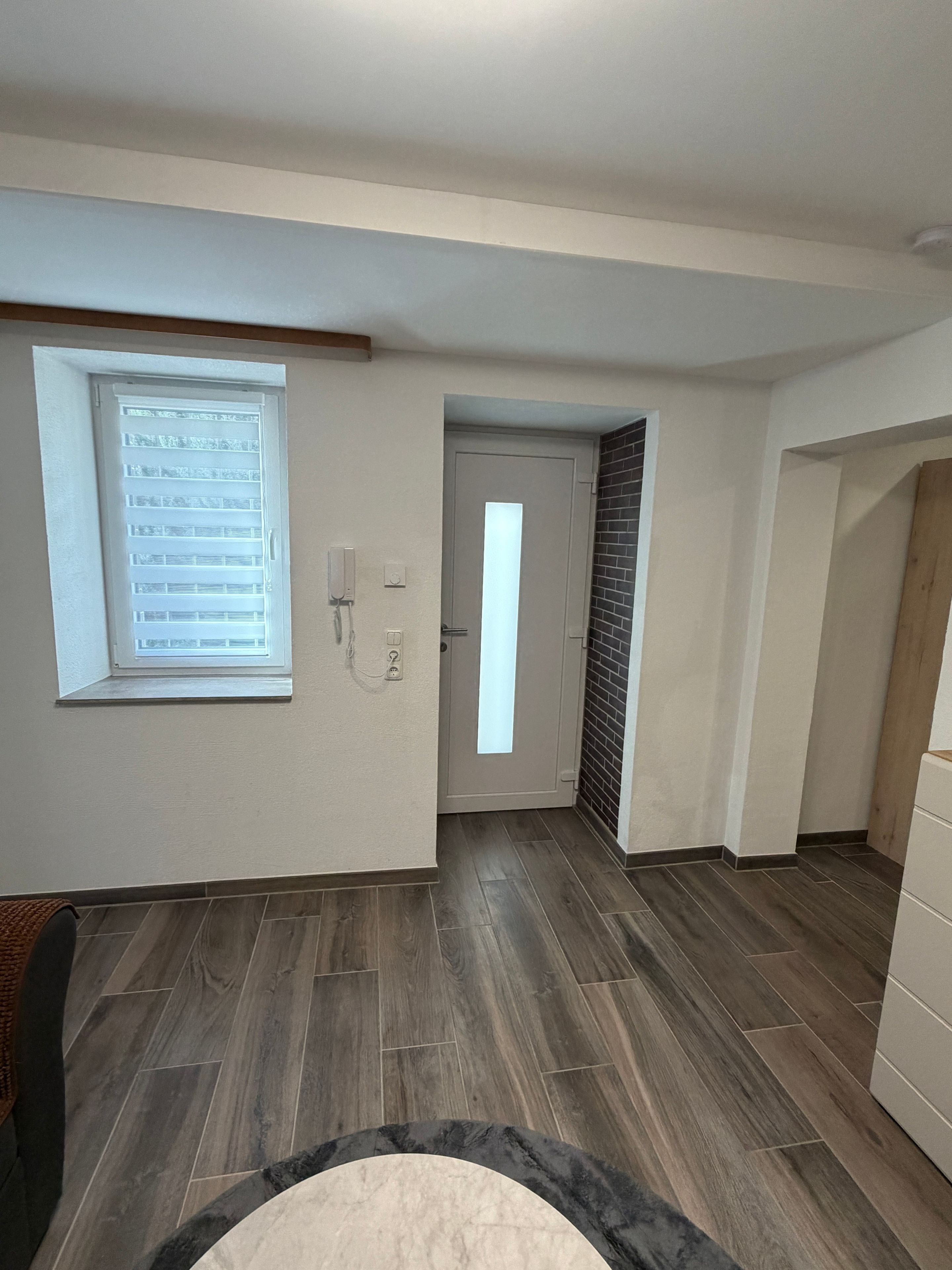 Ein Zimmer mit Holzboden, einem Fenster mit Jalousien, einer Tür und einem an der Wand befestigten Lichtschalter.
