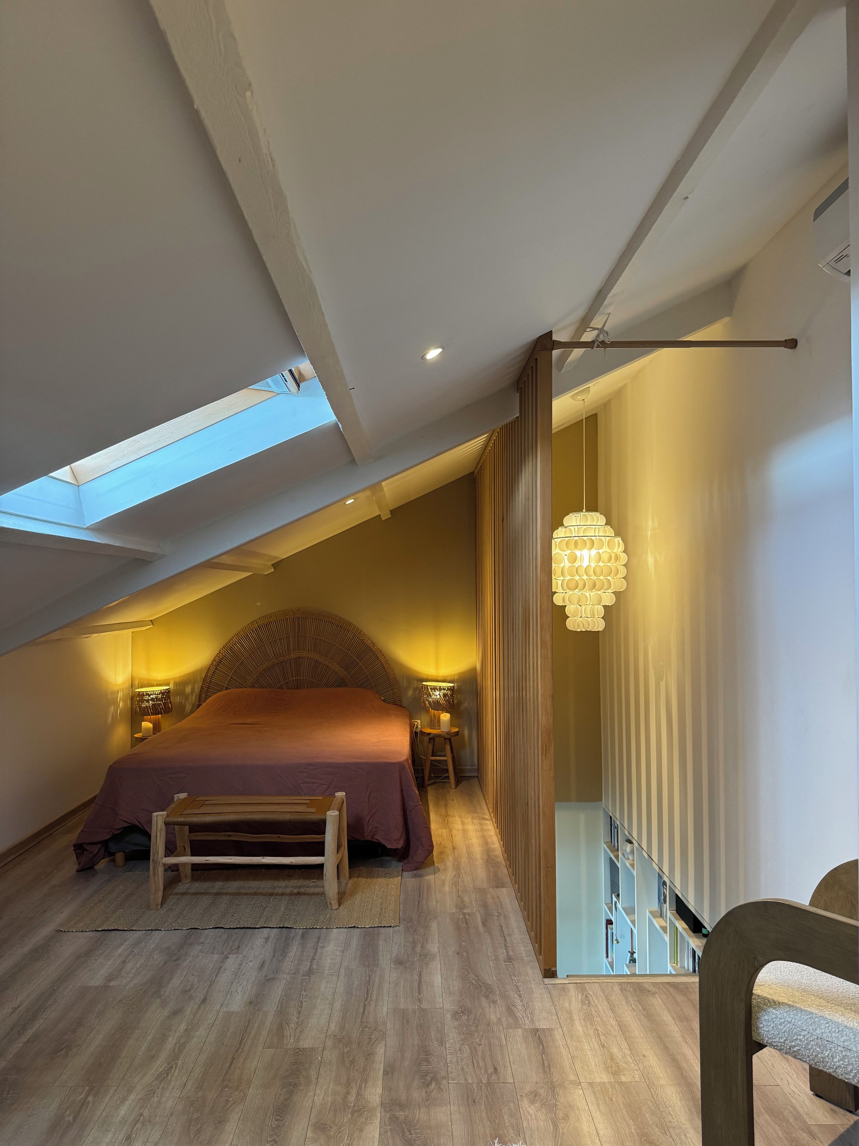 Une chambre avec un plafond en pente, un lit, un banc, un luminaire suspendu et un escalier.