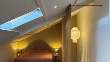 Une chambre avec un plafond en pente, un lit, un banc, un luminaire suspendu et un escalier.