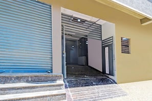 Entrada de um edifício com uma persiana azul fechada, escadas que levam a um andar superior e uma pequena janela com padrão de grade.