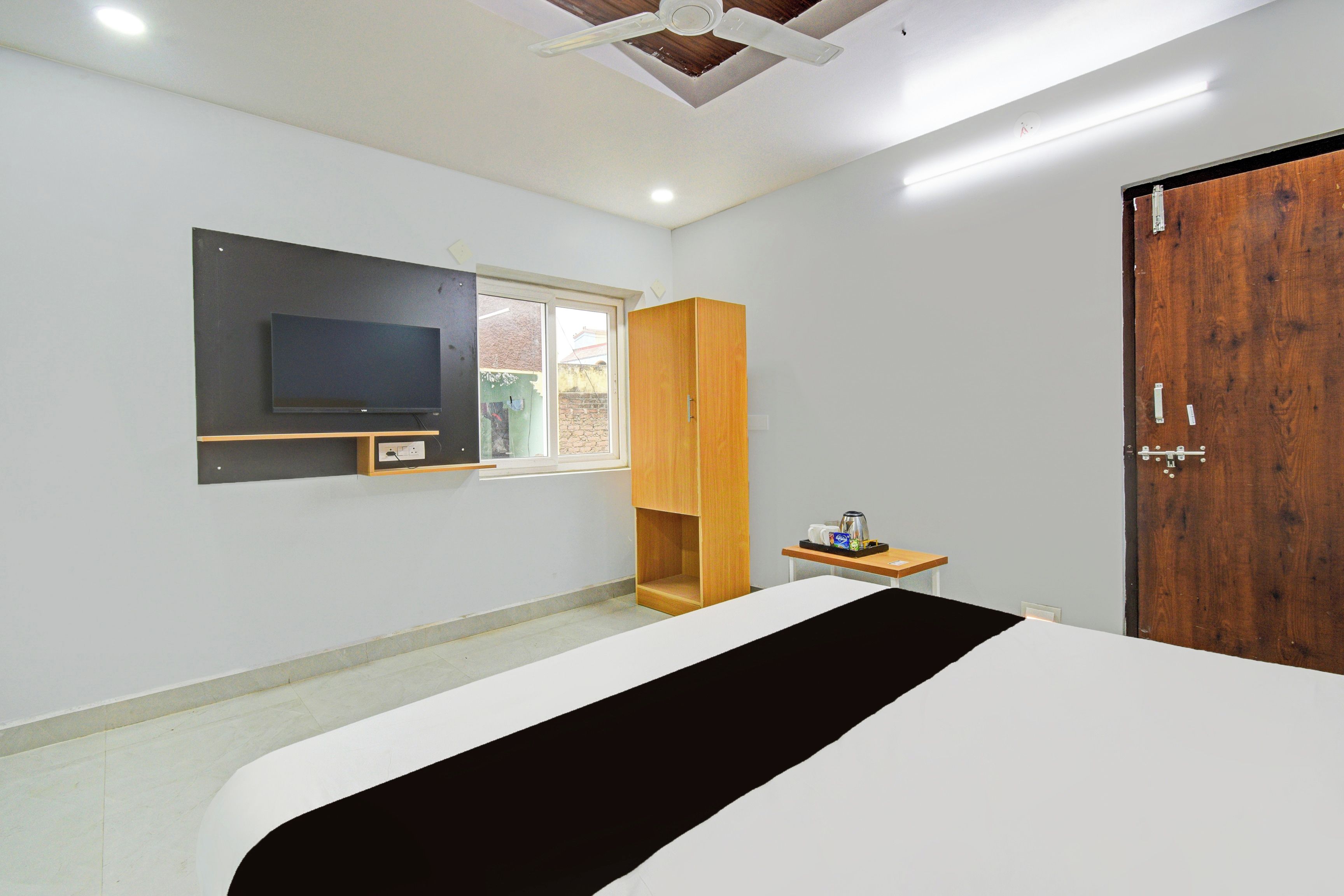 A bedroom with a bed, a television, a wooden wardrobe, a small table with a kettle, and a door.