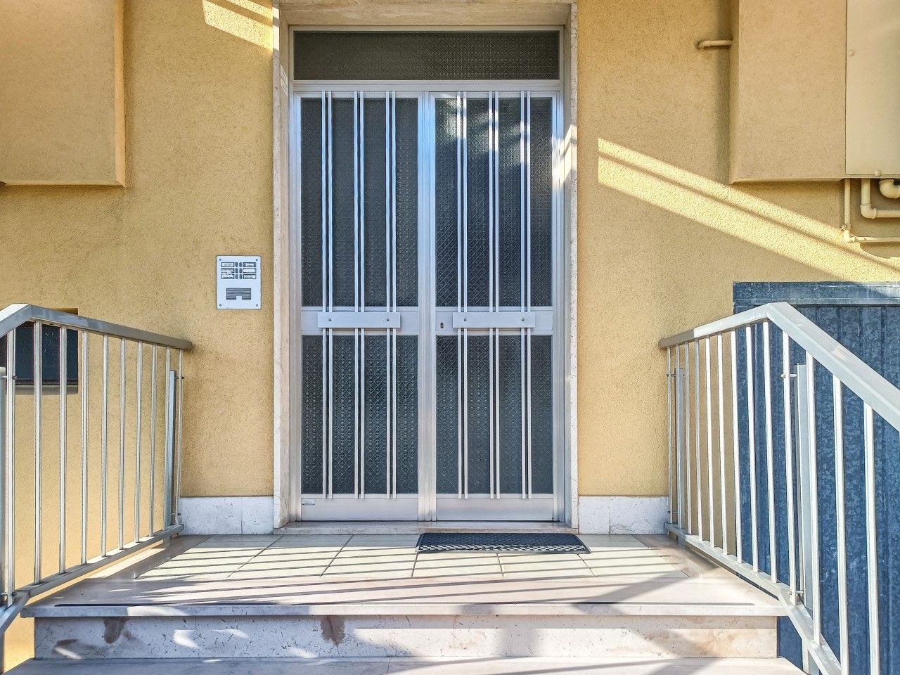 A building entrance with a glass door, metal railings, and a tiled ground.