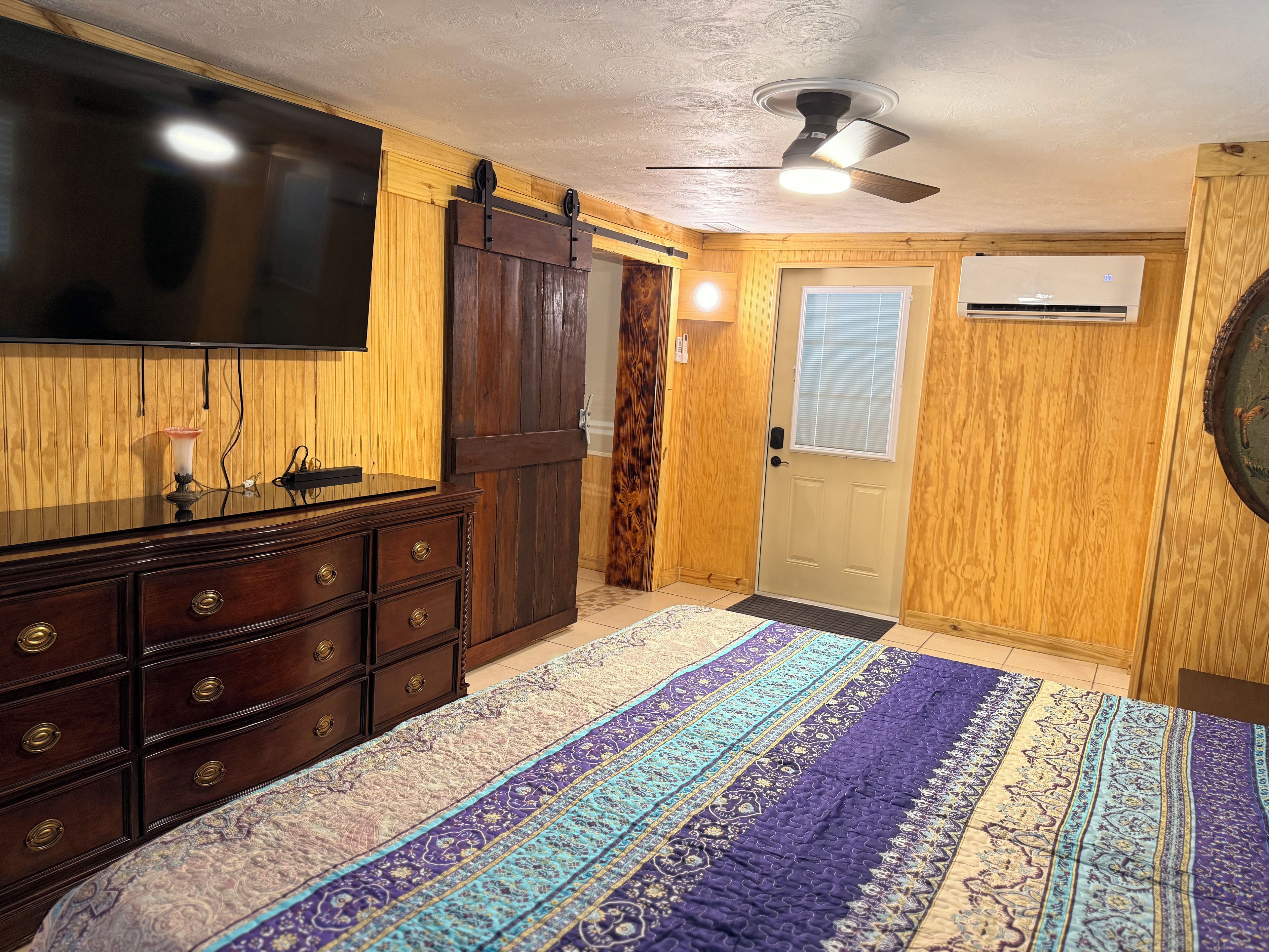 A bedroom with a bed, dresser, TV, ceiling fan, and an air conditioning unit.