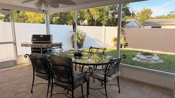 A covered patio with a glass table and four chairs, a ceiling fan, and a barbecue grill.