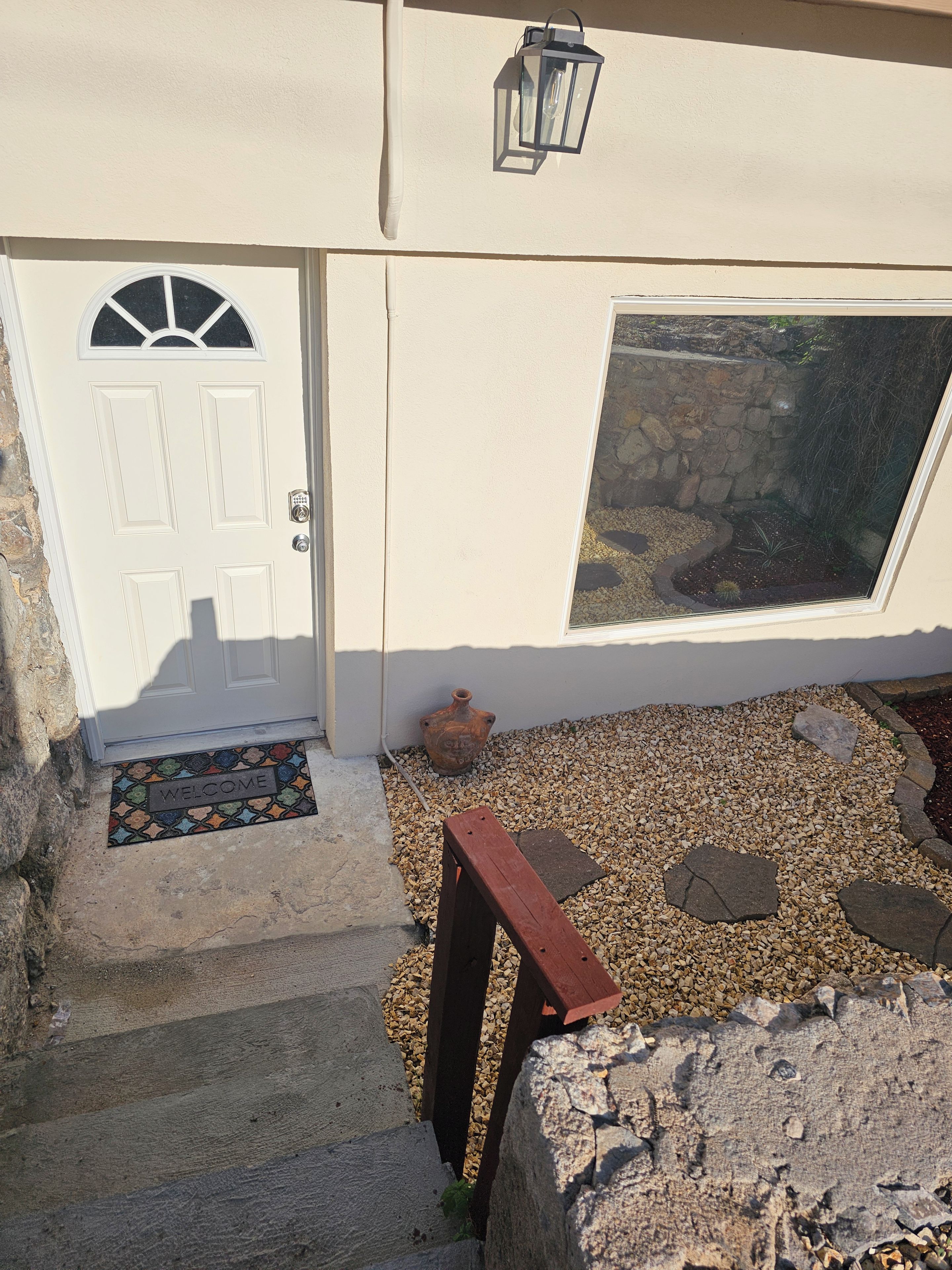 A white door with a window above it, a welcome mat, and a small garden with pebbles and a wooden bench.