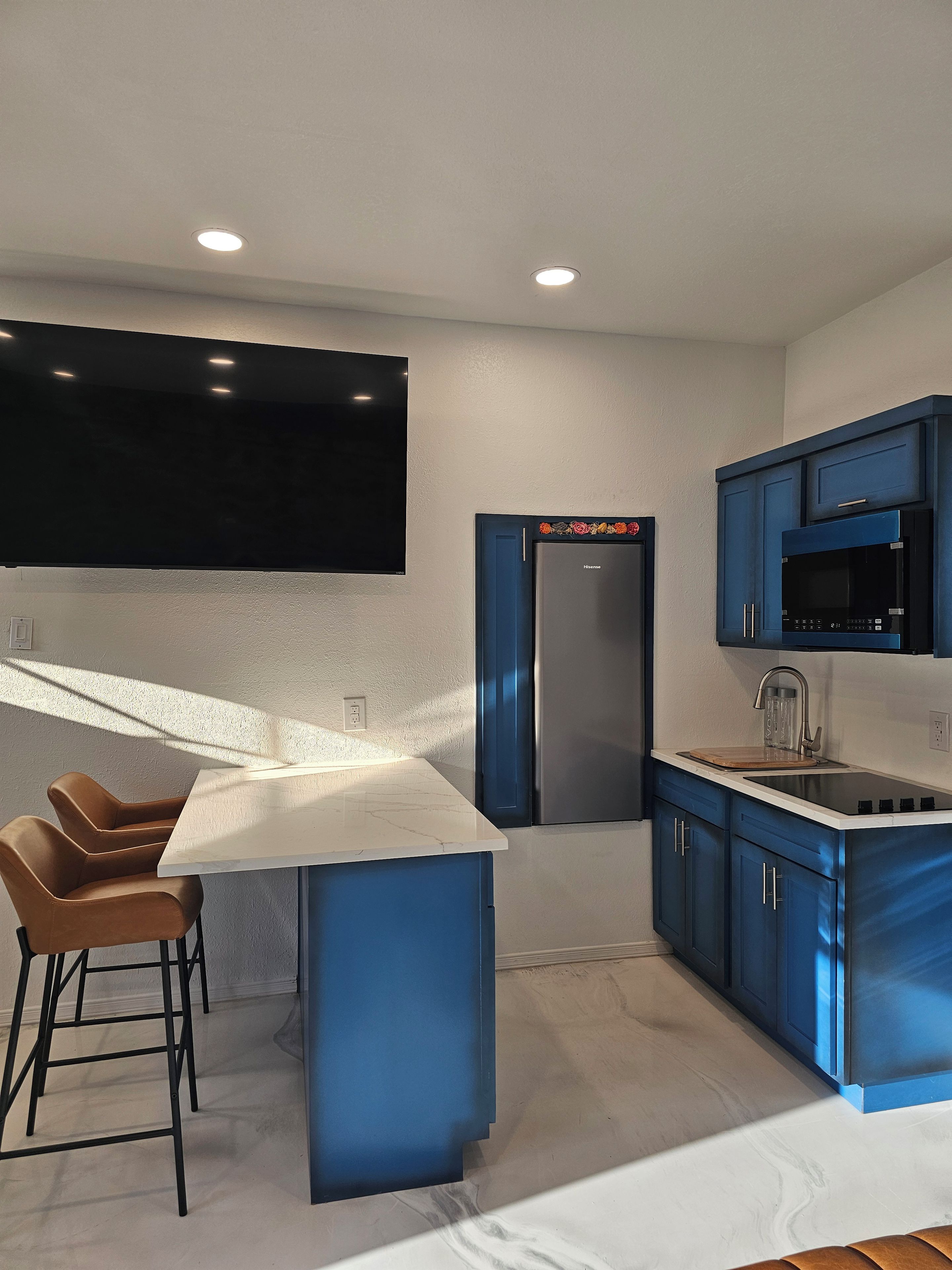 A modern kitchen with a central island, blue cabinets, a stainless steel refrigerator, and a flat-screen TV mounted on the wall.