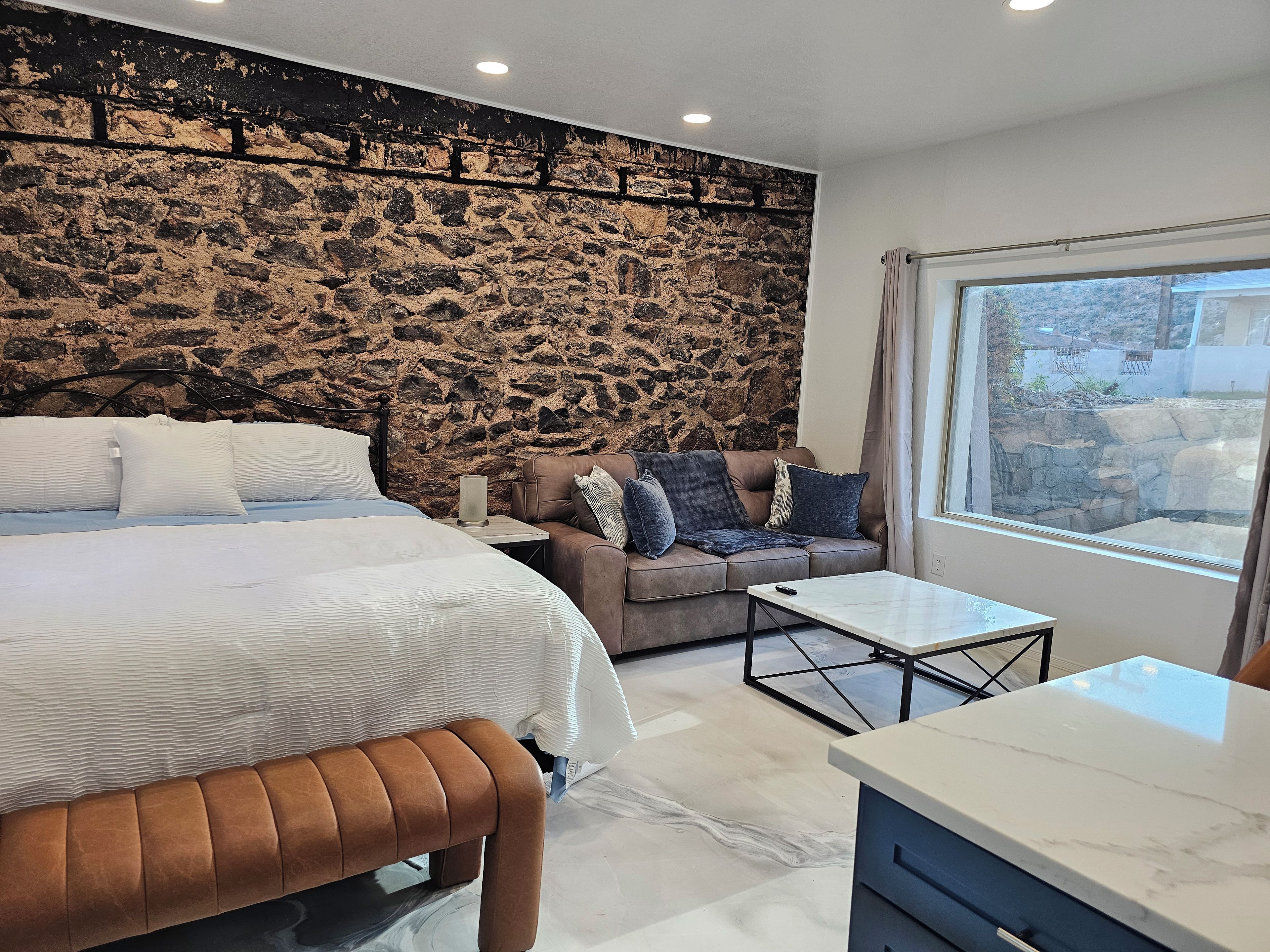 A modern bedroom with a stone wall, a bed, a sofa, and a view of the outdoors.