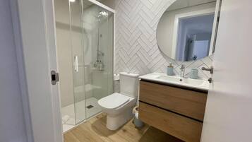 Un baño moderno con ducha envuelta en vidrio, inodoro blanco, tocador de madera con espejo y pared con estampado de espiga.