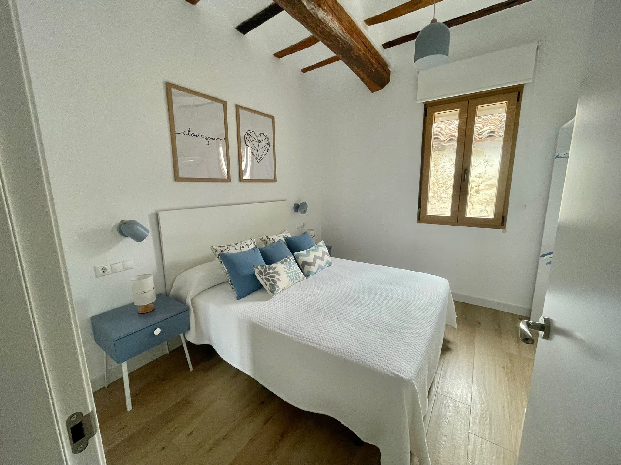 Un dormitorio con cama, mesitas de noche, ventana y techo con vigas a la vista.