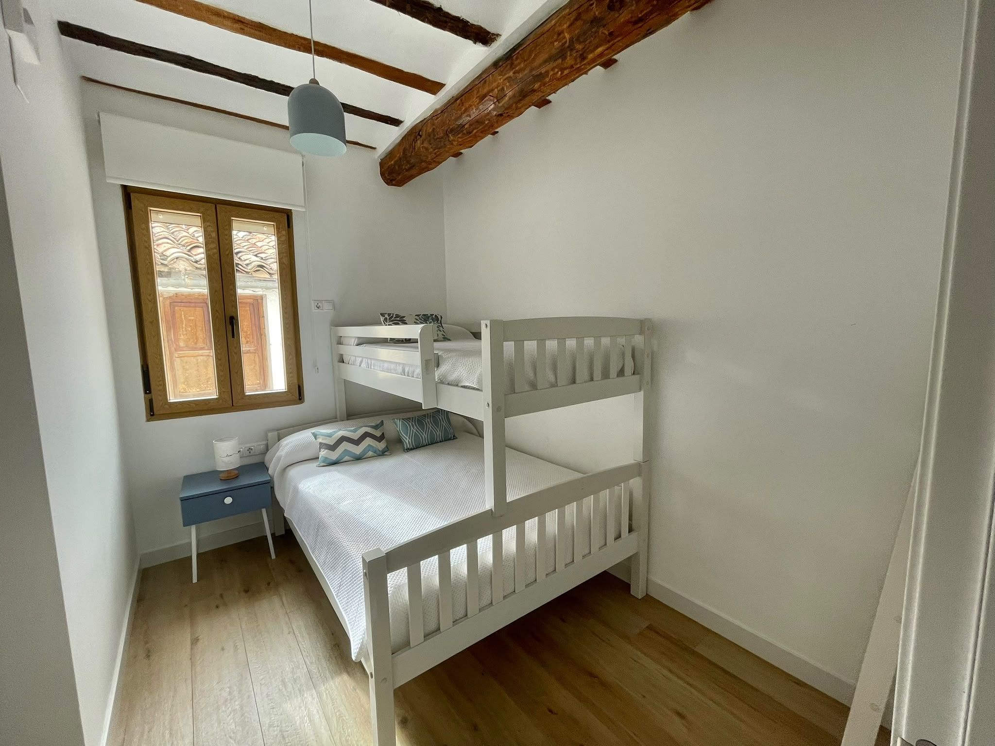 A small, single-bed room with a wooden bunk bed, a desk with a chair, a hanging light fixture, and a window with a view of the outside.