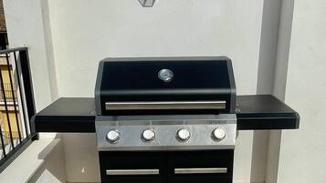 Una parrilla negra con cuatro quemadores y un estante lateral, ubicada en una azotea con un muro blanco y una barandilla metálica al fondo.
