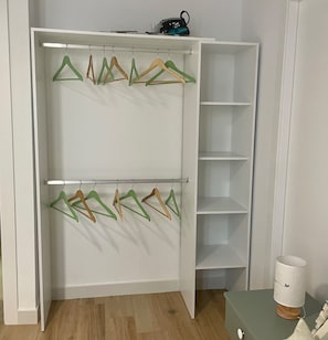 Une armoire blanche avec deux étagères et une tringle pour suspendre les vêtements, un plancher en bois et une petite table avec une lampe.