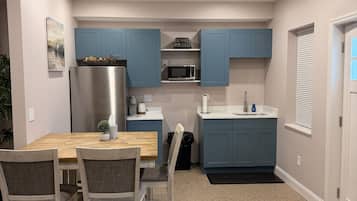 Een compacte keuken met blauwe kastjes, een roestvrijstalen koelkast, een magnetron en een eethoek met een houten tafel en stoelen.