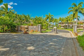 A paved entrance with a stone wall and a sign, palm trees, and a clear blue sky. - Haven Villa by Gather - 3BR Princeville Townhome w/ Anini Valley Views (Princeville)