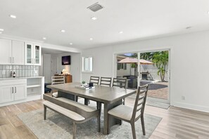 A modern kitchen with white cabinetry, a dining area with a wooden table and benches, and a sliding glass door leading to an outdoor patio. - Pool Home - Close to Barefoot & Bonita Beaches (Bonita Springs)