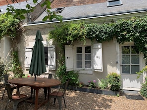 Une terrasse avec une table ronde en bois et des chaises, un parasol vert, et un bâtiment blanc doté de fenêtres et d’une porte.
