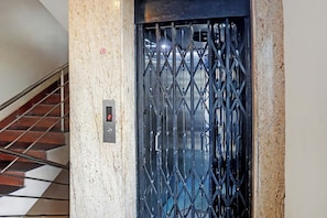 An elevator with a metal grille door, a control panel with a red light, and a staircase with a metal railing.