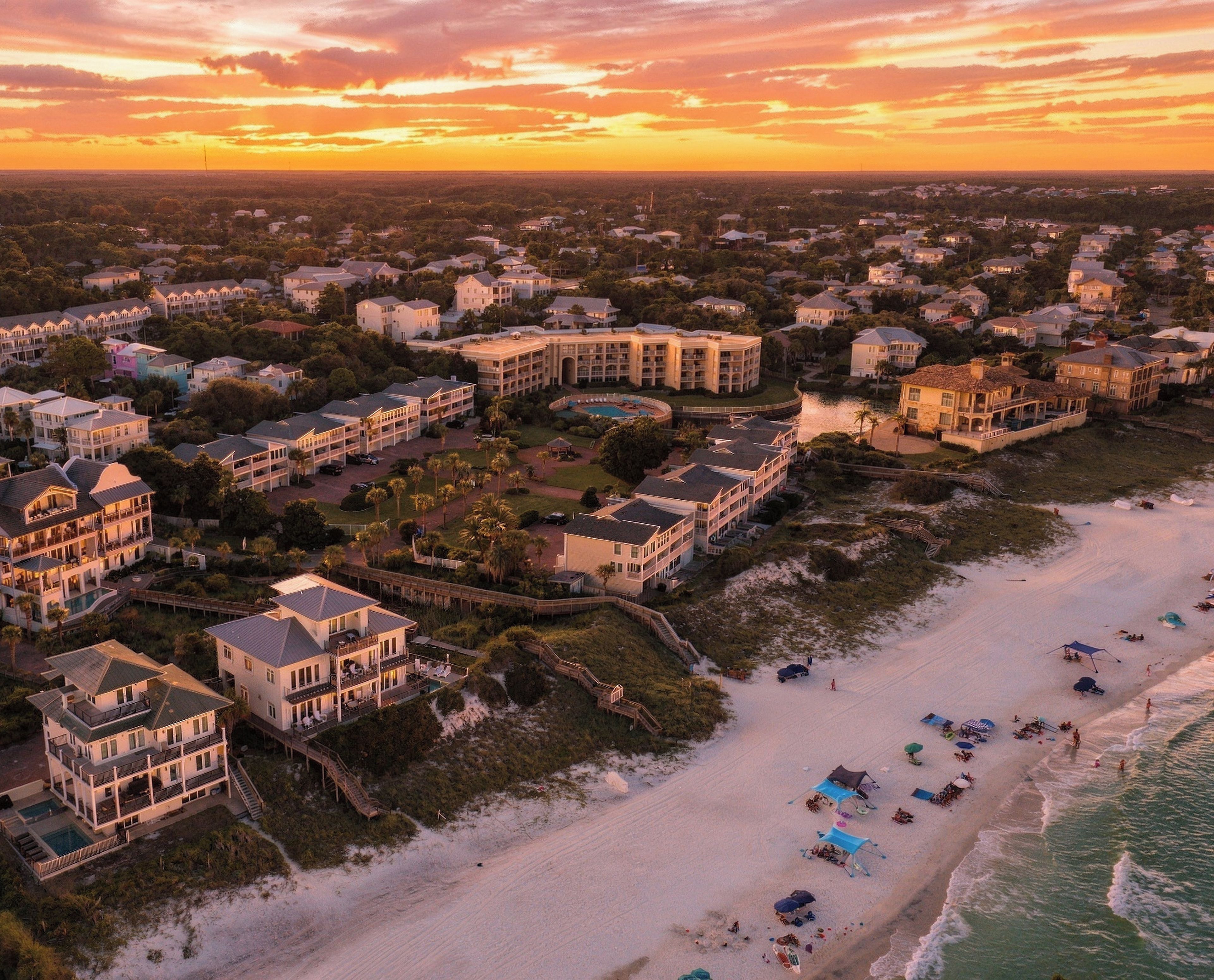 Aerial view of beachfront properties nestled along pristine white sand, with the surrounding coastal neighborhood visible under a dramatic sunset sky.