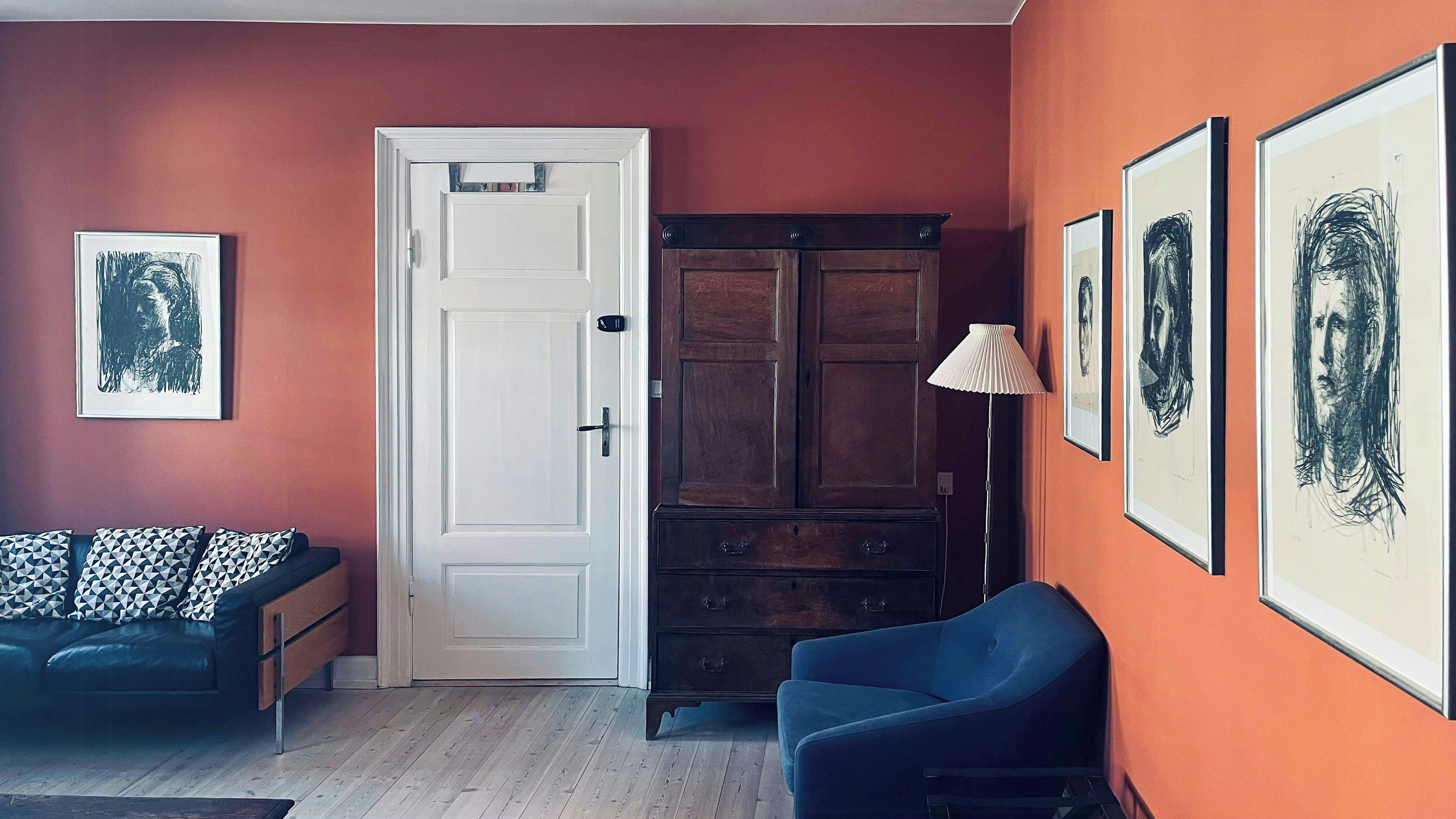 A room with a white door, a blue armchair, a dark wooden cabinet, and framed artwork on the walls.