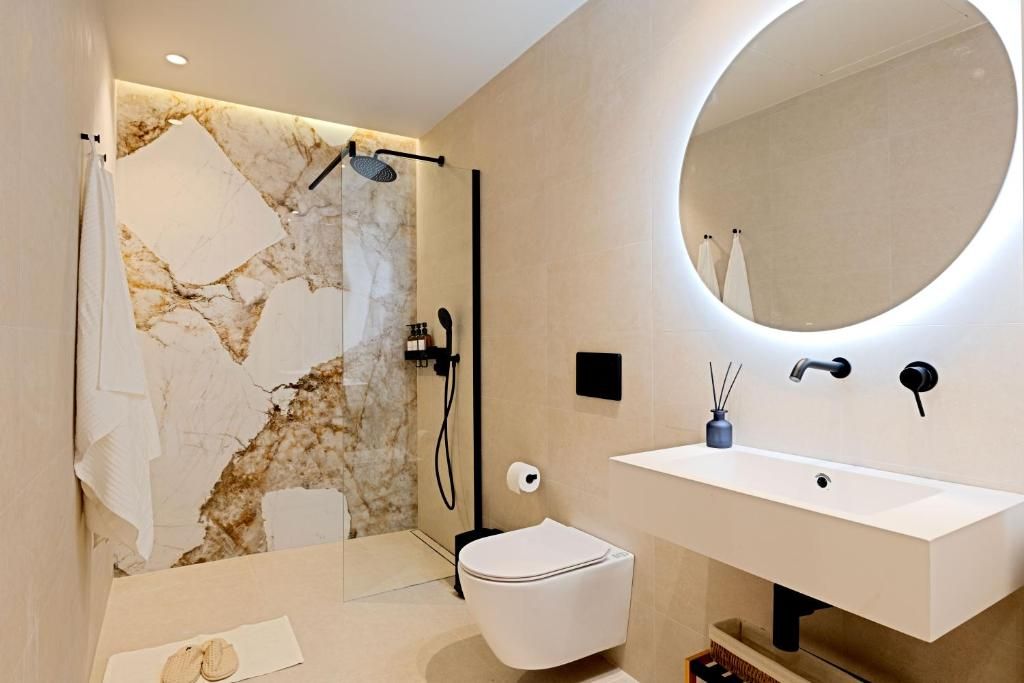 Un baño moderno con pared de mármol, un espejo redondo, un lavabo blanco y un inodoro.