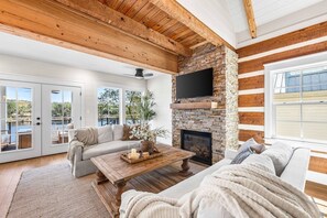 A spacious living room with a stone fireplace, a flat-screen TV, and a large window overlooking a deck. - Coastal Vista (Santa Rosa Beach)