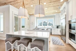 A modern kitchen with white cabinetry, a central island with stools, stainless steel appliances, and a view of buildings through large windows. - Amberjack (Panama City Beach)