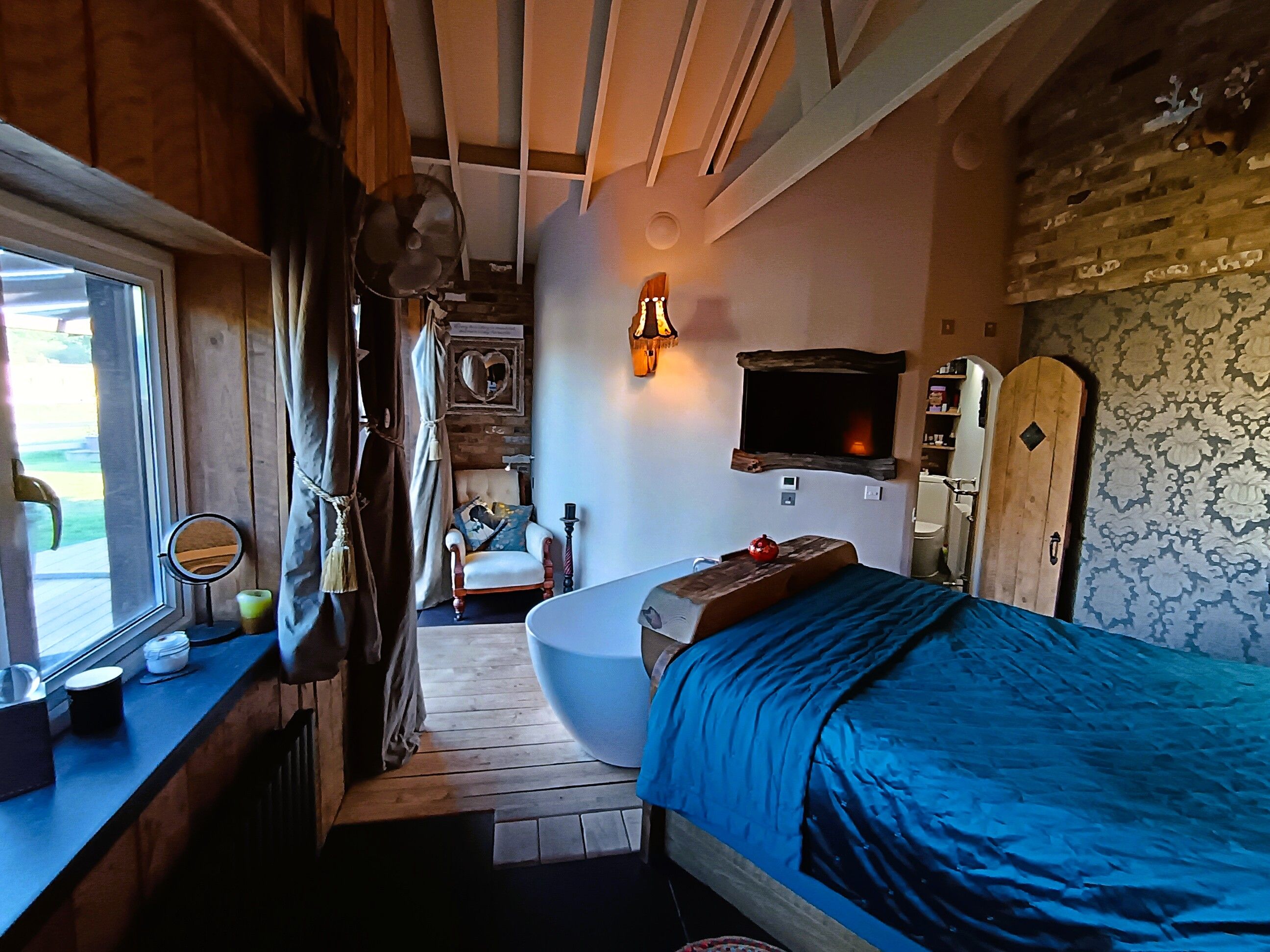 A compact bedroom with a single bed, a small bedside table, a mirror, and a window with curtains.