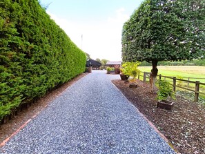 A gravel pathway leading to a building with a dark roof, surrounded by neatly trimmed hedges and a tree.