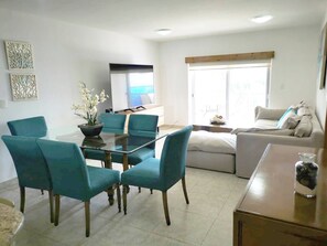 A modern living room with a glass dining table surrounded by teal chairs, a sofa, and a coffee table. - Wonderful Beachfront Condo in Cancun Hotel Zone (Cancún)