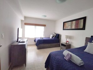 A hotel room with two beds, a desk, a chair, a window, and a painting on the wall. - Wonderful Beachfront Condo in Cancun Hotel Zone (Cancún)