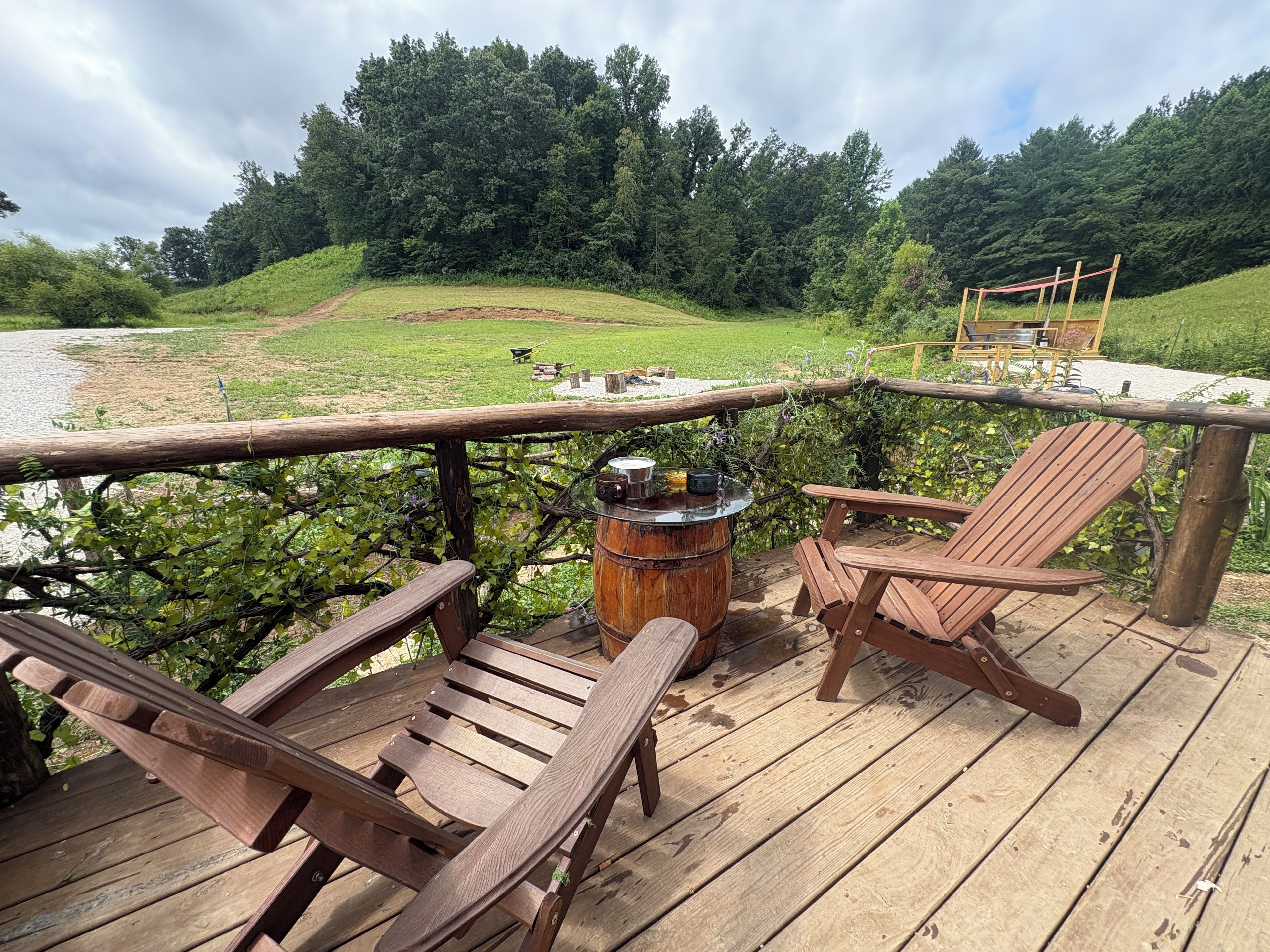 A wooden deck with two Adirondack chairs and a barrel, overlooking a grassy field and a forested hill.