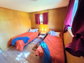Een kamer met twee bedden, elk met een blauwe deken, een klein tafeltje met twee lampen en een raam met gordijnen.
