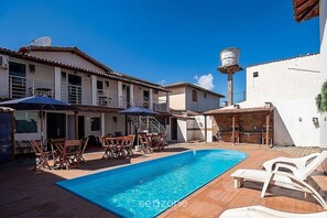 A swimming pool with lounge chairs and umbrellas, surrounded by buildings with balconies and a water tower. - Apt. in Condo. w/ Pool & BBQ Pax0003 (Porto Seguro)