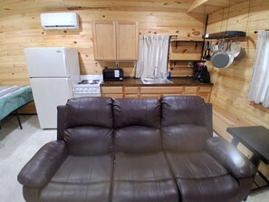 Una kitchenette compacta con refrigerador, estufa, microondas y ollas colgantes. Un sofá de cuero marrón está en primer plano.