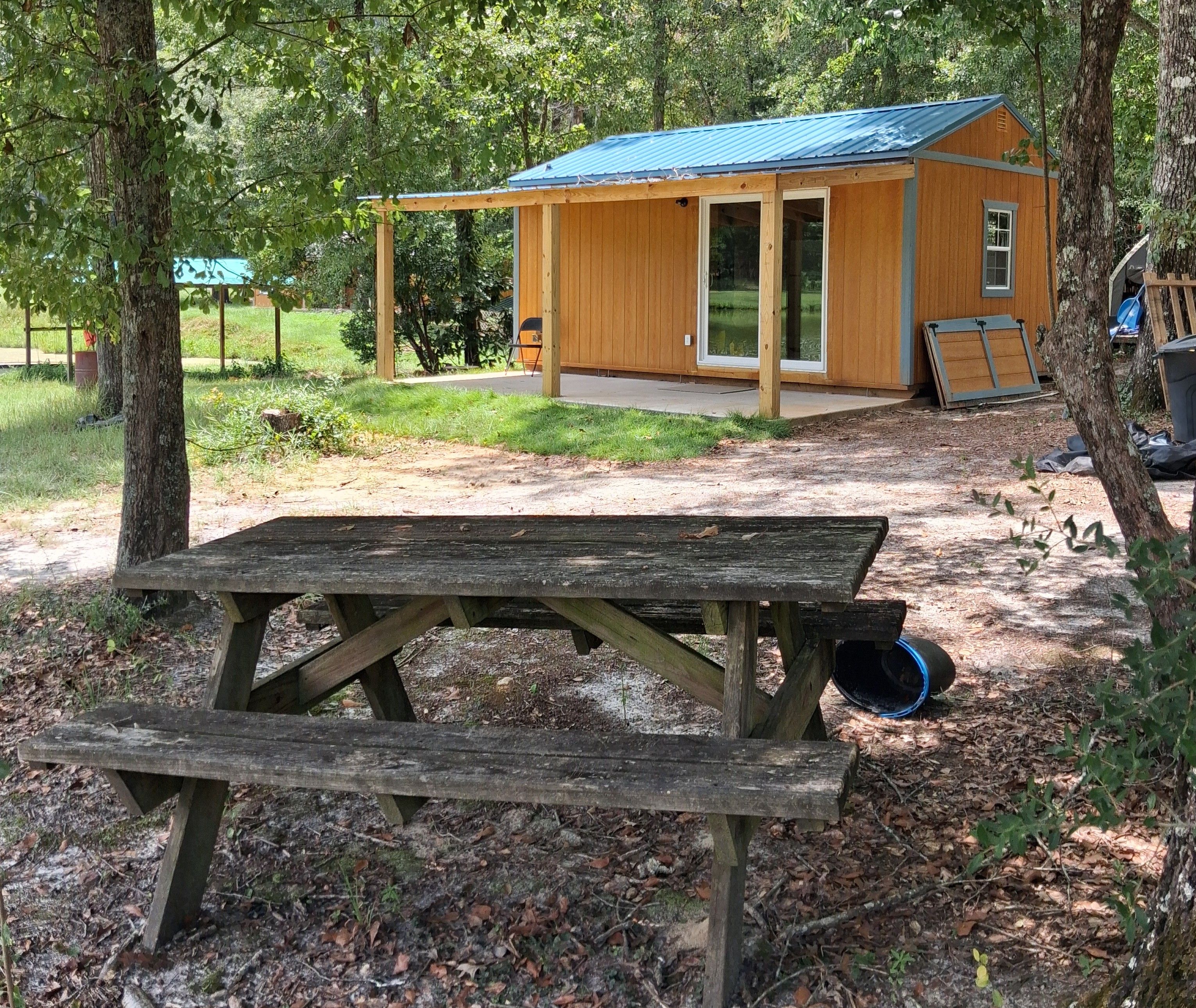 A wooden picnic table with a bench in a wooded area, a small wooden cabin with a blue roof, and a clear view of the surrounding trees.