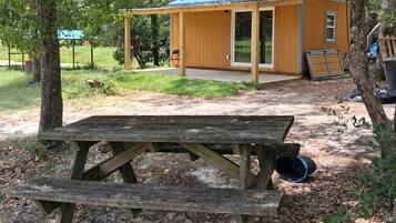 A wooden picnic table with a bench in a wooded area, a small wooden cabin with a blue roof, and a clear view of the surrounding trees.
