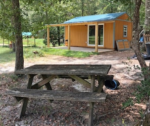 Una mesa de picnic de madera con banco en un área boscosa, una pequeña cabaña de madera con techo azul y una vista despejada de los árboles circundantes.