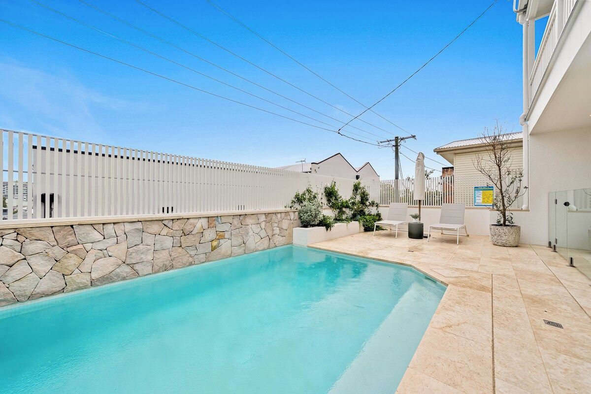 A swimming pool with a stone wall, a white fence, and a small patio area with a chair and a potted plant.
