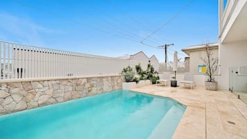 A swimming pool with a stone wall, a white fence, and a small patio area with a chair and a potted plant.