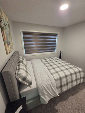 Une chambre à coucher avec un lit, une fenêtre avec des stores, une table de chevet et une œuvre d’art murale.
