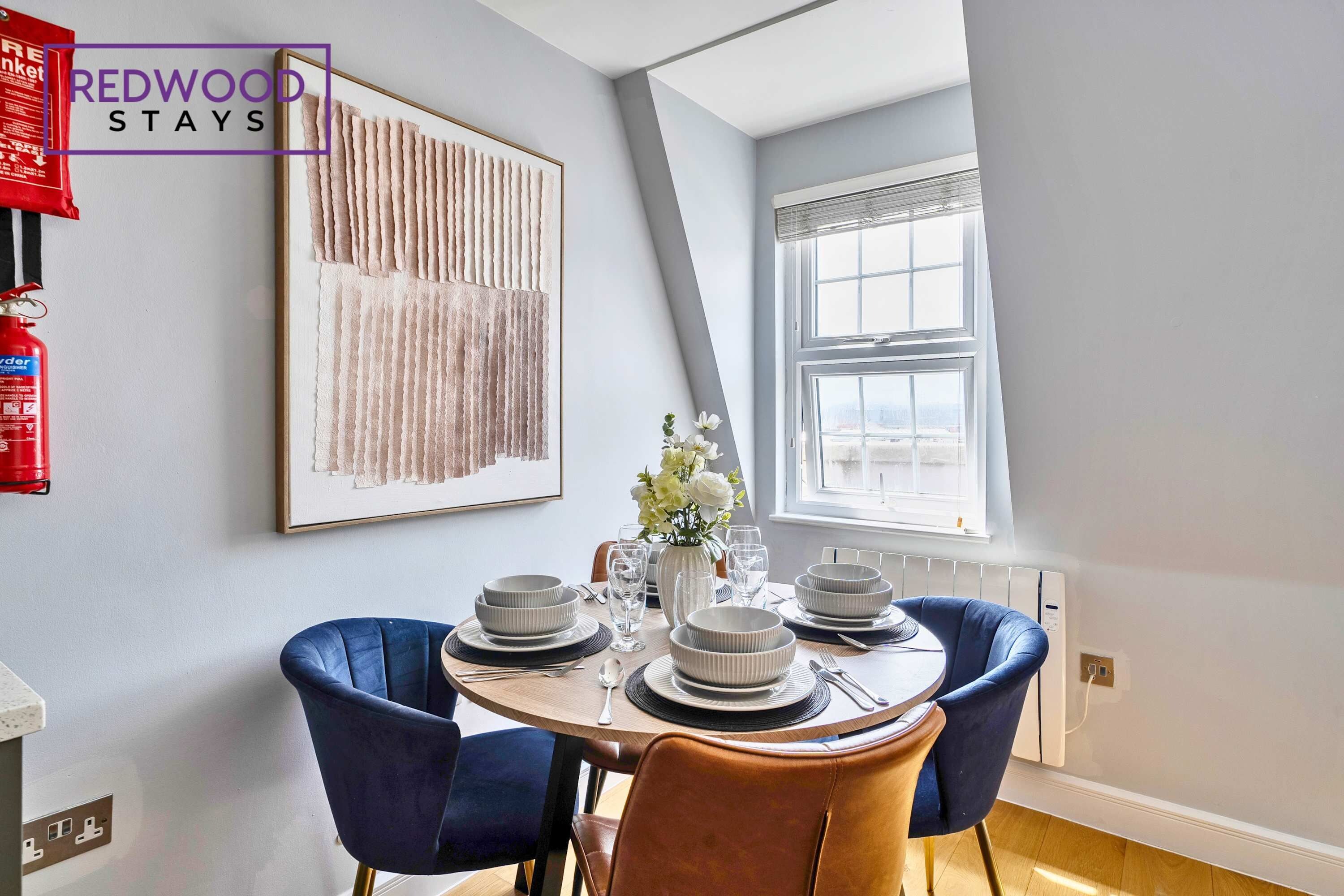A dining area with a round table, blue and brown chairs, white plates, and a vase with flowers. A framed artwork hangs on the wall, and a fire extinguisher is mounted on the wall.