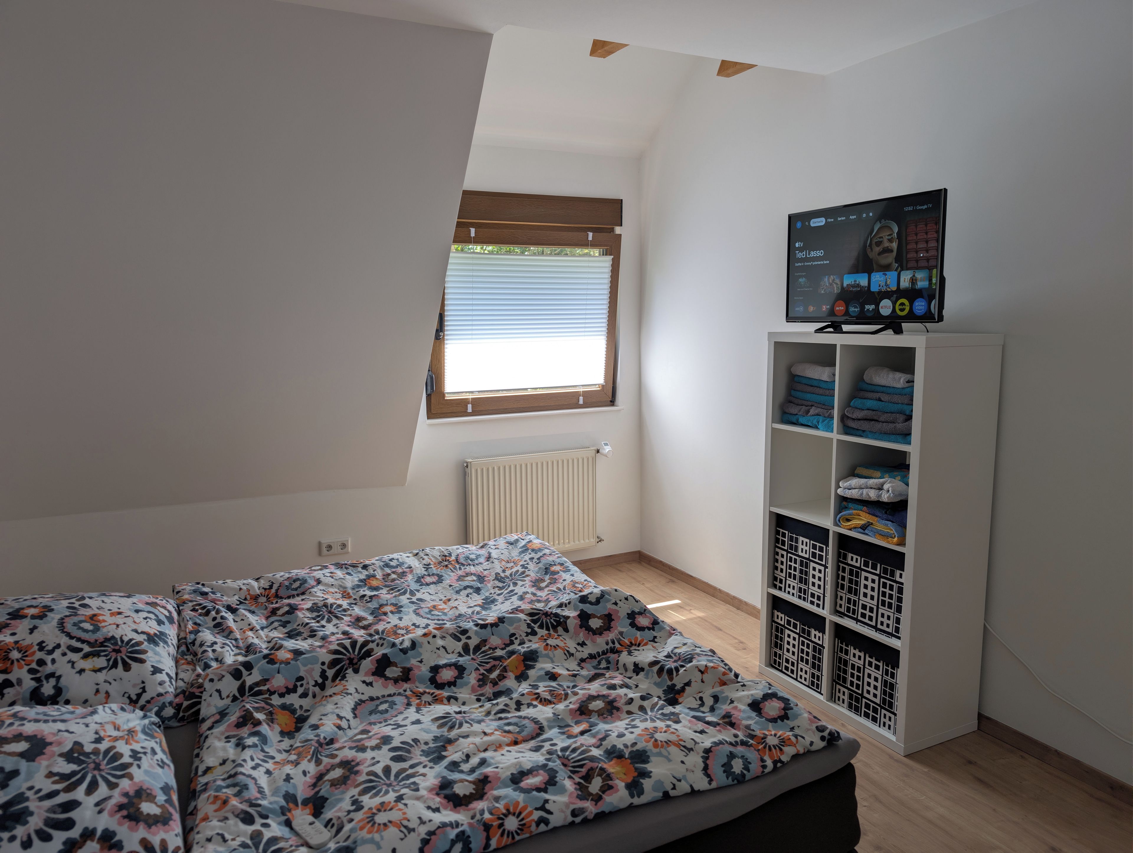 Un dormitorio con cama, ventana, radiador y televisor en un estante.