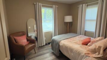 A bedroom with a bed, a chair, a bedside table, a lamp, and a window with curtains.