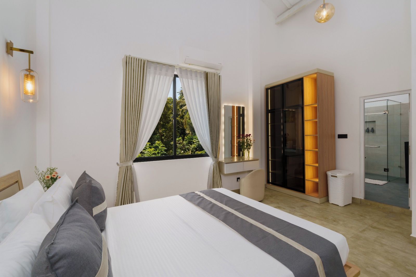 Ein modernes Hotelzimmer mit einem großen Bett, einem Schminktisch mit Spiegel, einem begehbaren Kleiderschrank und einer Glasdusche.
