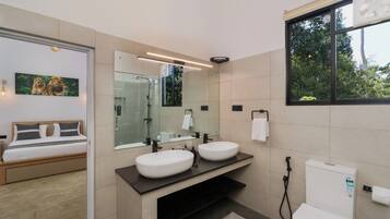 Une salle de bain moderne avec un double lavabo, une douche et des toilettes. La pièce adjacente contient un lit avec une tête de lit.