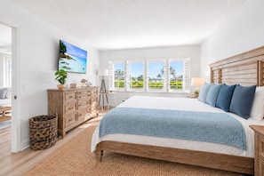 Une chambre avec un grand lit, une commode en bois, un tableau et une vue sur la verdure par la fenêtre.