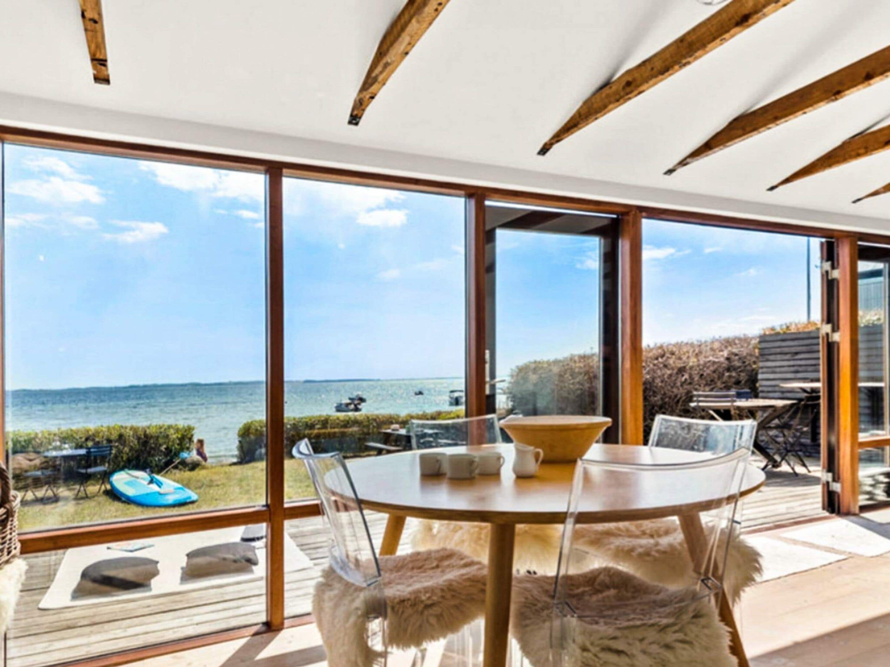 A modern dining area with a glass table and chairs, large windows offering a view of the sea, and wooden beams on the ceiling.