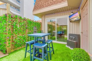 Un patio con una mesa y sillas, una cerca de madera con plantas trepadoras y un ventanal corredizo que da a un jardín.